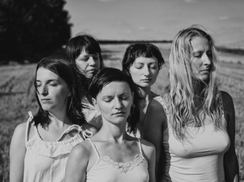 Czarno-biała fotografia przedstawia grupę pięciu młodych kobiet. Kobiety są ubrane w letnie sukienki, stoją w otwartej przestrzeni na polu, mają zamknięte oczy. Oświetla je mocne słońce.