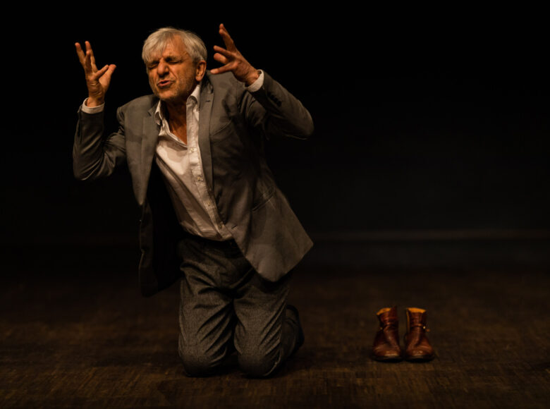 Na ciemnej scenie klęczy siwiejący mężczyzna w średnim wieku. Jest ubrany w ciemny garnitur i jasną koszulę. Ma zaciśnięte oczy i grymas na twarzy, a jego ręce uniesione są do góry w bardzo ekspresyjnym geście. Obok niego na podłodze stoi para męskich butów.