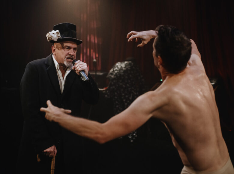 Zdjęcie ze spektaklu. Na scenie znajduje się półnagi mężczyzna. Na przeciwko niego stoi mężczyzna w czarnym fraku i kapeluszu. Otaczają ich kotary.