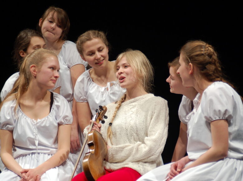 Zdjęcie ze spektaklu młodzieżowego. Na czarnym tle widać siedem dziewcząt. Sześć z nich jest ubranych w takie same białe sukienki. Włosy mają zaplecione w warkocze lub podpięte. W centralnym punkcie siedzi dziewczyna z jasnym warkoczem. Jest ubrana w beżowy sweter i czerwone spodnie, w rękach trzyma skrzypce. Pozostałe dziewczęta są skupione wokół niej, pochylają się do niej, słuchają jej.