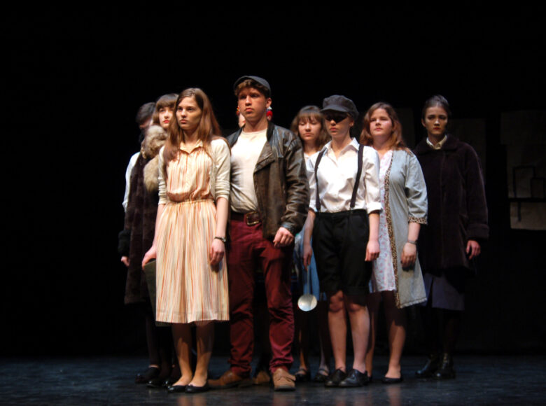 Zdjęcie ze spektaklu młodzieżowego. Na czarnej podłodze i na czarnym tle grupa młodych osób - dziewczęta i chłopcy. Ubrani są w stroje nawiązujące do czasów wojennych. Jest dziewczyna w futrze, dziewczyna w jasnej sukience poniżej kolan, chłopak w bordowych spodniach, skórzanej kurtce i czapce kaszkietówce. Stoją jakby tworzyli mur, wszyscy patrzą na wprost, w jeden punkt.