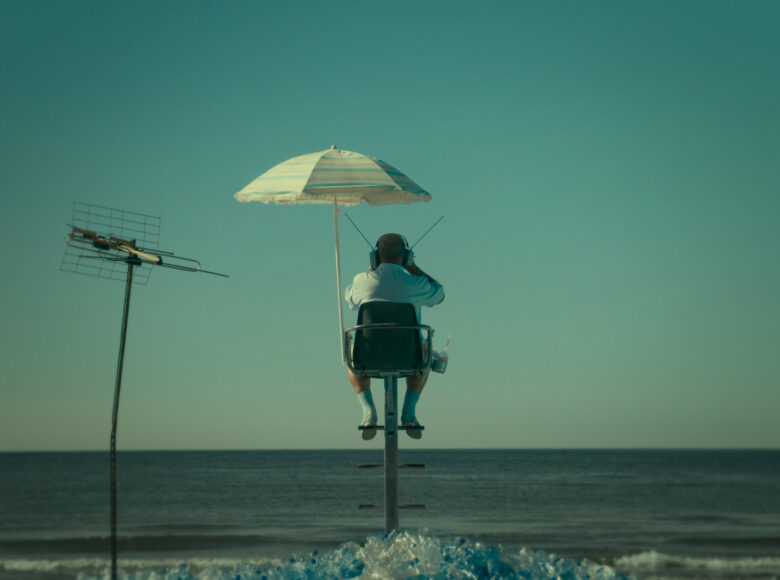 Kadr z filmu. Na brzegu morza, na krześle ratownika, pod parasolem siedzi mężczyzna. Widzimy go od tyłu. Na głowie ma duże słuchawki, każda z anteną. Na pierwszym planie góra plastikowych butelek i stojąca na brzegu antena.
