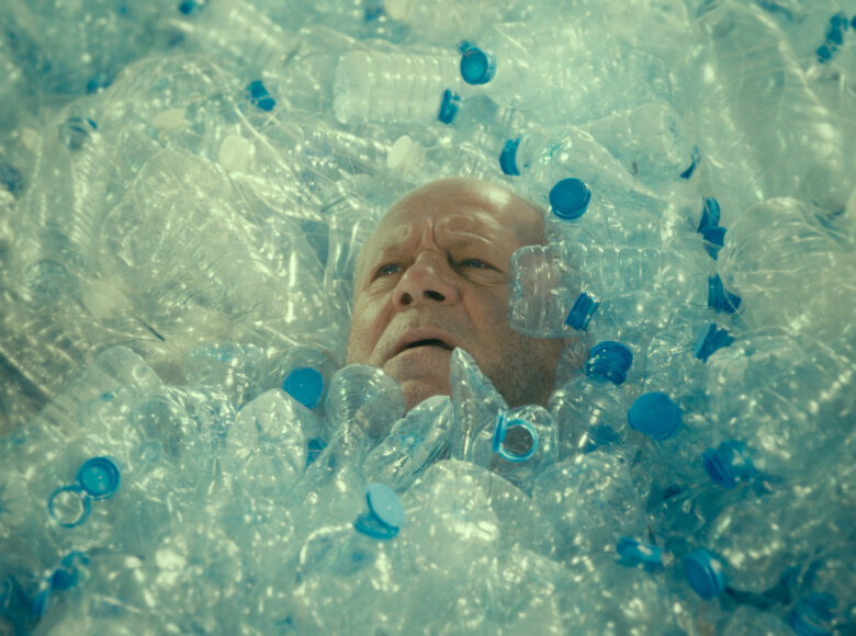 Kadr z filmu. Twarz starszego mężczyzny otoczona mnóstwem pustych, plastikowych butelek z niebieskimi nakrętkami.