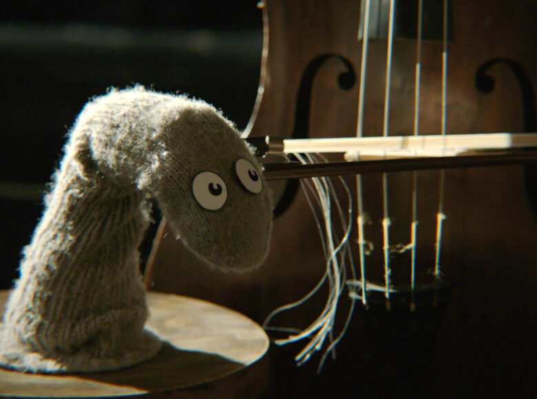 Kadr z filmu. Na okrągłym blacie stoi skarpetka z oczami. Trzyma smyczek i gra na wiolonczeli.
