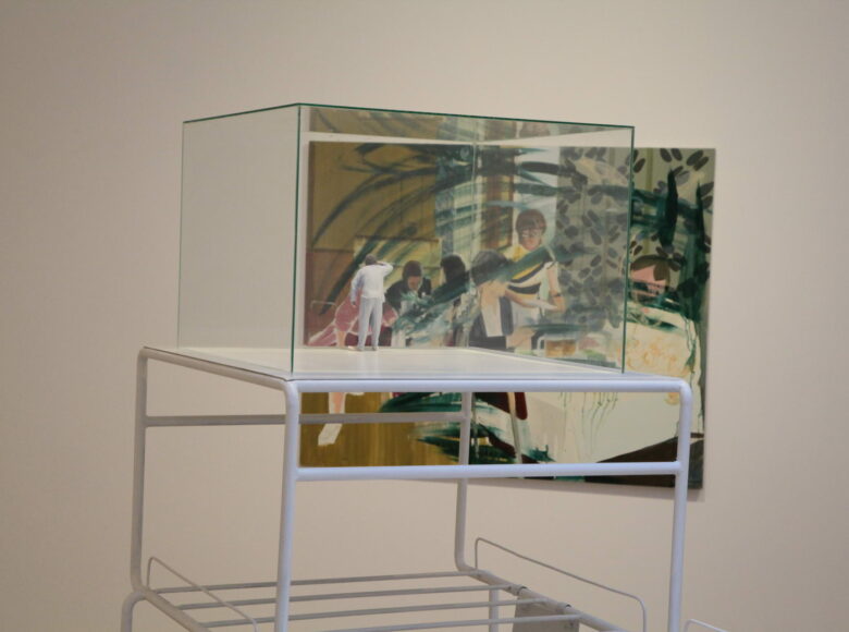 Zdjęcie przedstawia artystyczną instalację składającą się ze szklanego, sześciennego naczynia. W środku naczynia znajduje się figurka mężczyzny.