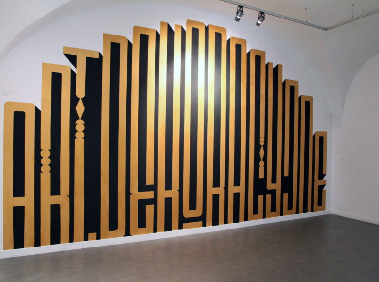 Zdjęcie przedstawia graffiti na białej ścianie. To wielkie, złote litery w stylu art deco układające się w napis: art. dekoracyjne.