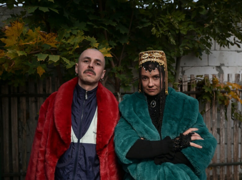 Zdjecie. Męzczyzna i kobieta w kolorowych futrach stoją w ogrodzie i patrzą przed siebie. Za nimi znajduje się drewniany płot i klon.