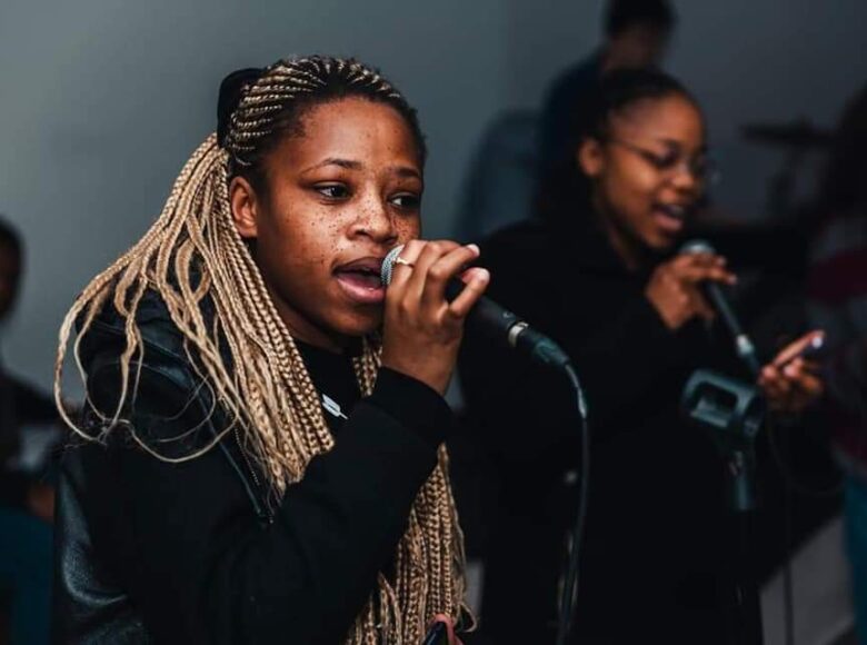 Zdjęcie przedstawia czarno-skórą dziewczynę, w czarnej bluzie i z jasnymi warkoczykami. Dziewczyna trzyma w ręce mikrofon i śpiewa.