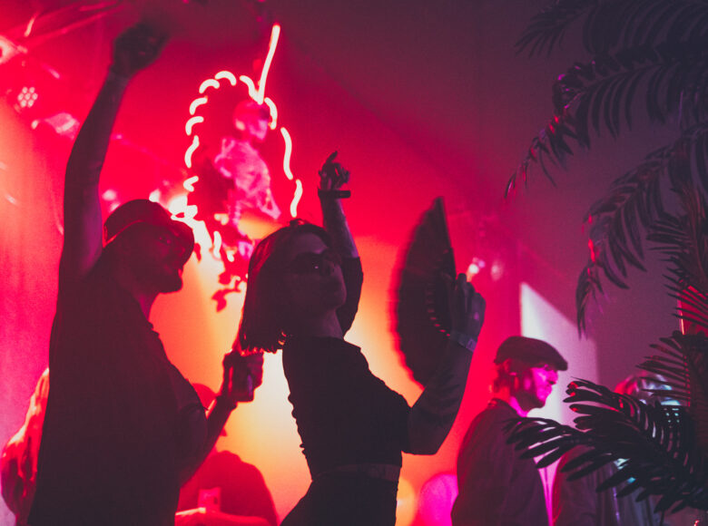 Zdjęcie przedstawia ludzi podczas imprezy. Na parkiet pada czerwone światło. Na ścianach świeca neony. Młoda kobieta tańczy, w ręce trzyma wachlarz.