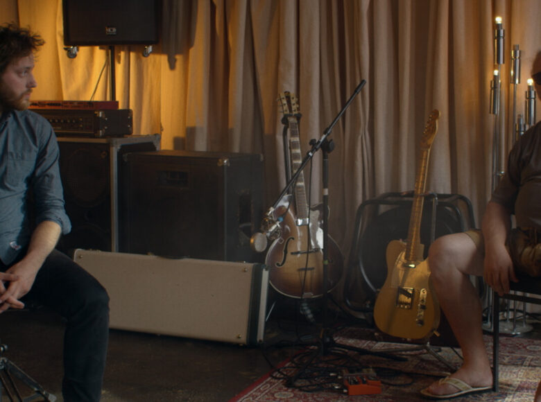 Kadr z filmu. Dwóch mężczyzn siedzi w studio nagraniowym. W tle stoją gitary, wzmacniacze i głośniki. Po lewej siedzi młody mężczyzna w ciemnej koszuli i spodniach, patrzy na starszego mężczyznę siedzącego po prawej. Ten jest w krótkich sporniach, klapkach, ma siwą brodę i długie, związane włosy, ciemne okulary.