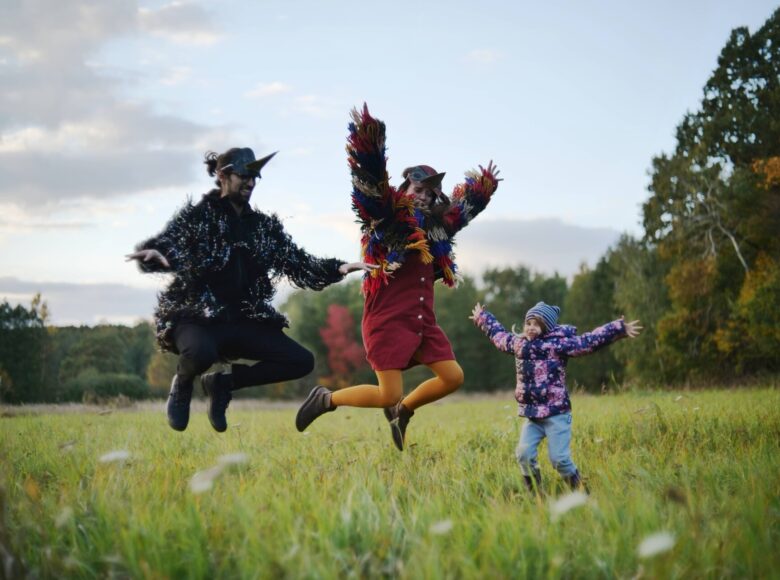 Zdjęcie przedstawia dwoje dorosłych ludzi i dziecko, znajdujących się na łące. Kobieta i mężczyzna przebrani w kostiumy ptaków skaczą. Dziecko stoi obok i wymachuje rękami.