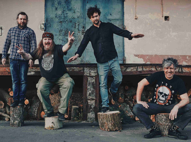 Zdjęcie przedstawia czterech mężczyzn, którzy siedzą albo stoją na drewnianych pieńkach. Mężczyźni uśmiechają się, są rozbawieni.