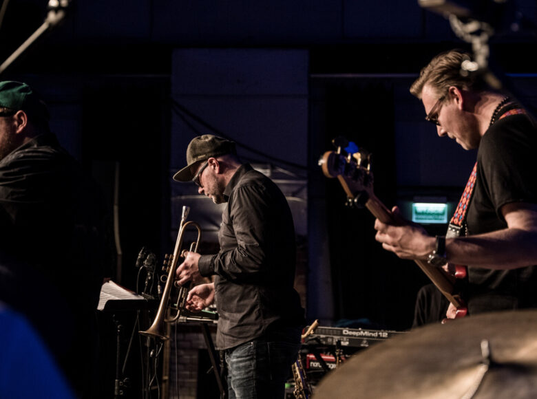 Zdjęcie przedstawia muzyków na scenie podczas koncertu. Jeden śpiewa, drugi gra na trąbce a trzeci na gitarze elektrycznej.