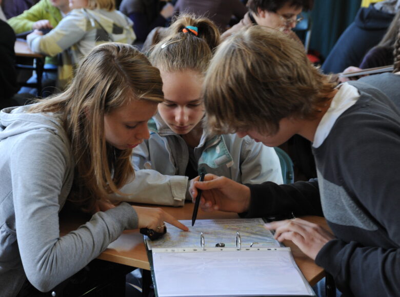 Zdjęcie przedstawia grupę młodych ludzi, która siedzi przy stoliku i pochyla się nad otworzonym papierowym segregatorem.