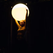 Na ciemnej scenie stoi kobieta i wyciąga nad głowę ręce, w których trzyma podświetlana, biała piłkę.