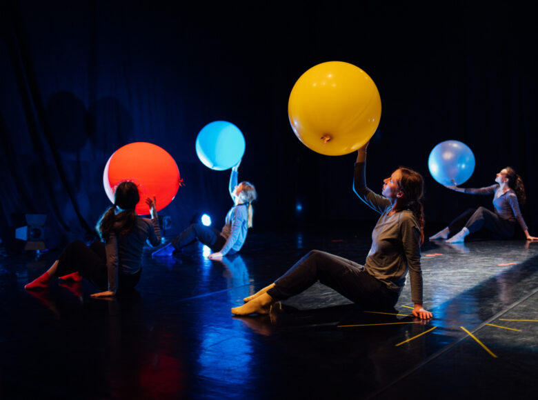 Zdjęcie przedstawia kilka kobiet, które siedzą na scenie. W jednej ręce unoszą nad siebie kolorowe, podświetlane piłki i na nie patrzą.