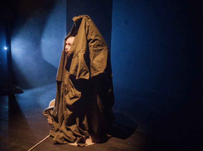 Zdjęcie ze spektaklu "Afryka Kazika". Na scenę pada niebieskie śiwatło. Z namiotu wykonanego z brązowego materiału wystaje głowa mężczyzny.