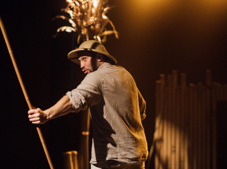 Zdjęcie ze spektaklu "Afryka Kazika". Mężczyzna w jasnym ubraniu i podrózniczym kapeluszu stoi na scenie. W ręce trzyma pałkę z trzciny. Za nim znajduje się palma i szałas z bambusowych palików.