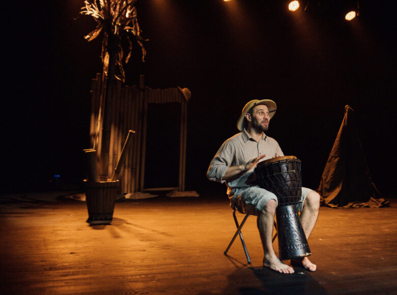 Zdjęcie ze spektaklu "Afryka Kazika". Bosy mężczyzna siedzi na taborecie i gra na bębnie.