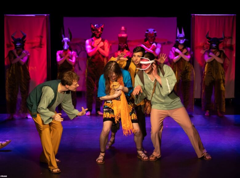 Zdjęcie ze spektaklu. Na scenie stoi obok siebie grupa aktorów i wykonuje ekspresyjne gesty. Pod jedną ze ścian stoi kilka osób przebranych za egipskie posagi z maskami byków na głowach.