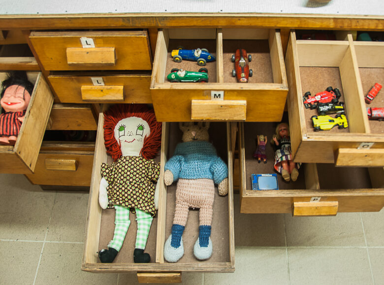 Zdjęcie przedstawia kilka drewnianych, otwartych szuflad. W szufladach znajdują się małe zabawki: samochodziki, laleczki i szmaciane maskotki.