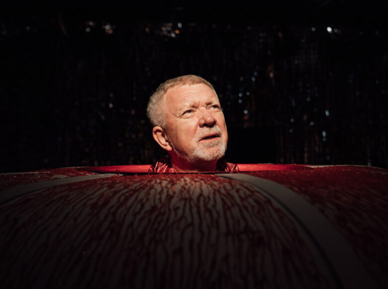 Zdjęcie ze spektaklu "Komedia Judyty". Przedstawia głowę starszego mężczyzny. Głowa wyłania się z metalowej konstrukcji, pokrytej czerwonymi wstążkami imitującymi krew.