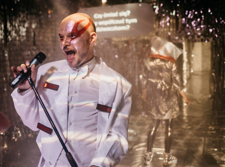 Zdjęcie ze spektaklu "Komedia Judyty". Aktor w biały stroju i makijazem na twarzy ma szeroko otworzone osta, w rękach trzyma statyw z mikrofonem. Podłoga i ściany wokół niego są wyłożone srebrną, błyszczącą folią.