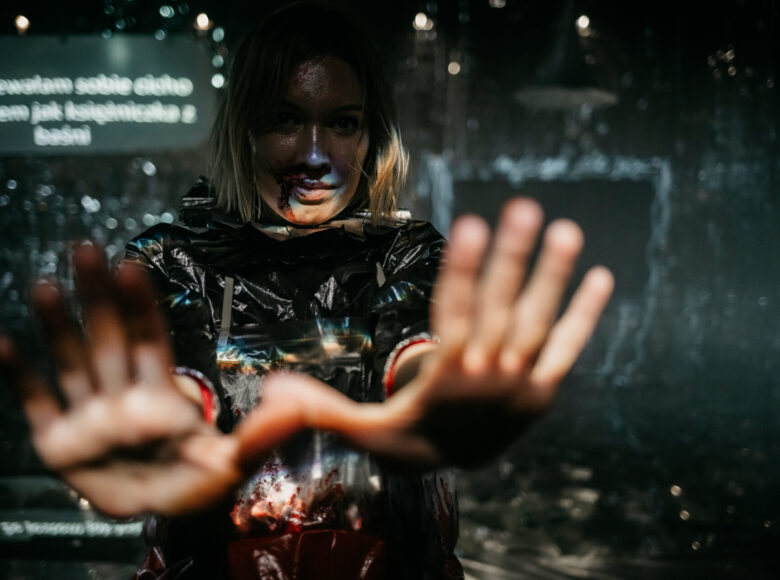 Zdjęcie ze spektaklu "Komedia Judyty". Na pierwszym planie znajduje się kobieta w srebrnym kostiumie. Kobieta wyciąga przed siebie obie ręce. Na jej kostiumie i twarzy znajduje się krew. Ściany i podłoga pomieszczenia wyłożone są srebrnym, błyszczącym materiałem.