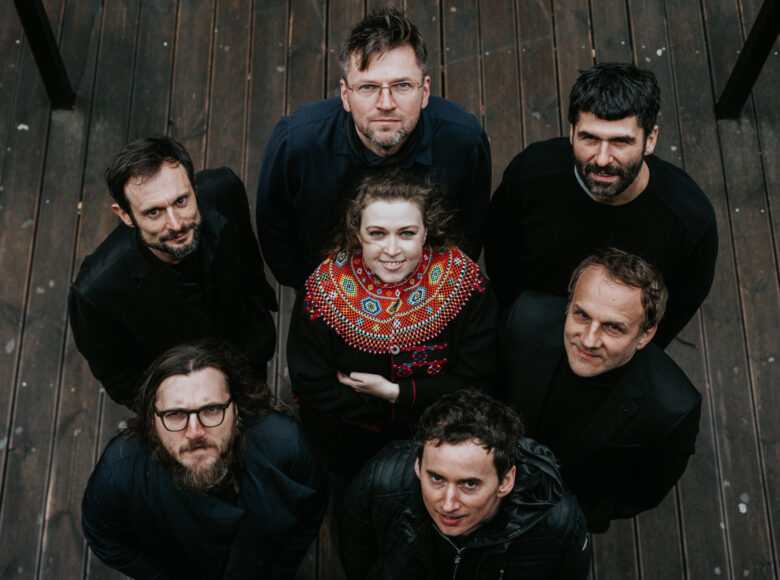 Zdjęcie przedstawia grupę muzyków z zespołu Tołhaje. Mężczyźni są ubrani na czarno i stoją w kręgu. W środku kręgu znajduje się kobieta w stroju z etnicznym haftem na kołnierzu. Grupa stoi na drewnianej pomoście. Patrzą w górę.