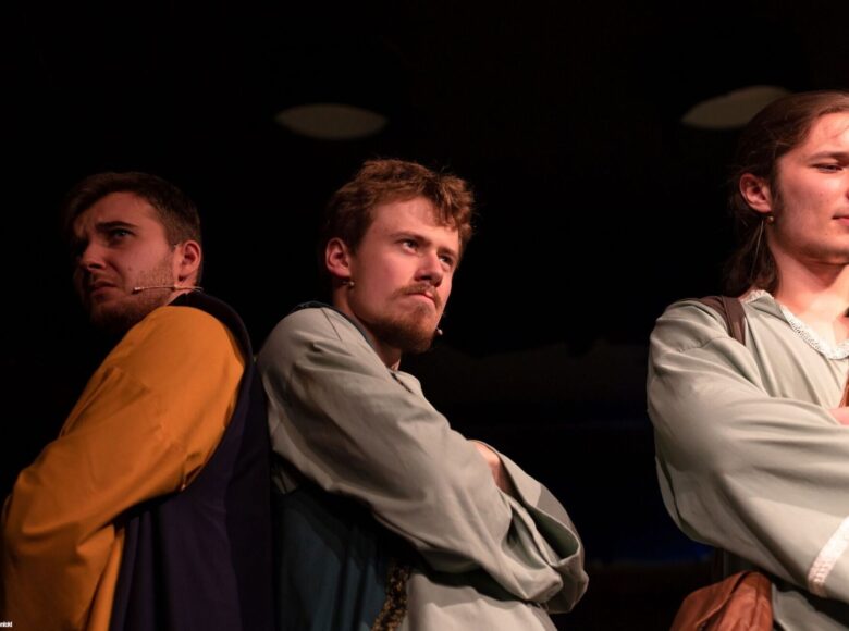 Zdjęcie ze spektaklu. Przedstawia trzech młodych mężczyzn w lnianych koszulach i kamizelkach. Stoją obok siebie, mają poważne, zamyślone miny.