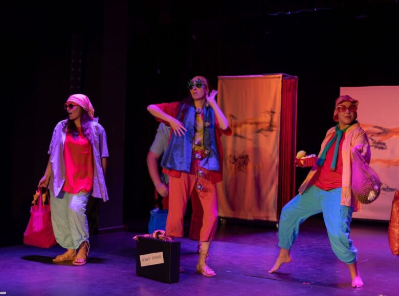 Zdjęcie ze spektaklu. Na scenie stoją trzy dziewczyny w letnich kolorowych strojach. Za nimi stoją białe parawany.