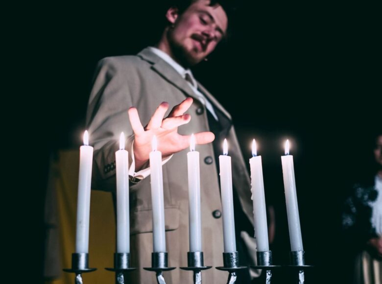 Zdjęcie ze spektaklu. Mężczyzna w garniturze zbliża rękę do metalowego świecznika, na którym pala się białe świeczki.