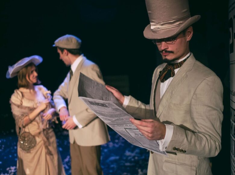 Zdjęcie ze spektaklu. Mężczyzna w kremowym garniturze i cylindrze na głowie stoi obok słupa ogłoszeniowego i czyta gazetę. Za nim znajduje się para w strojach z epoki, która patrzy sobie w oczy.