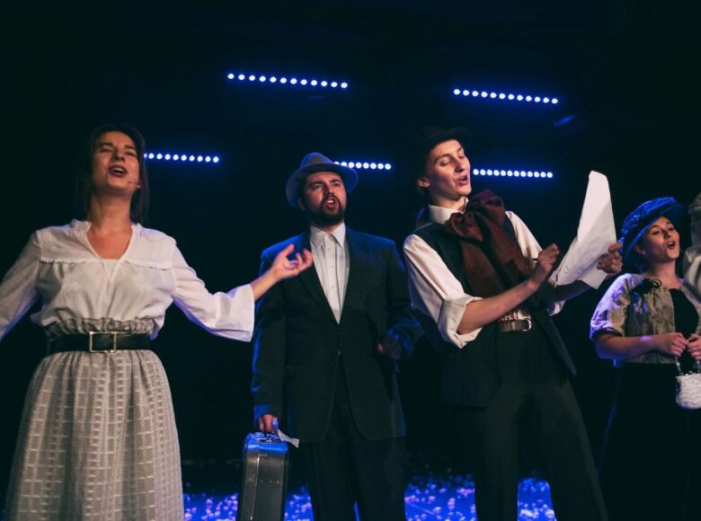 Zdjęcie ze spektaklu. Na scenie, w rzędzie stoi obok siebie grupa aktorów. Śpiewają. Za nimi świeca niebieskie neonowe lampki.