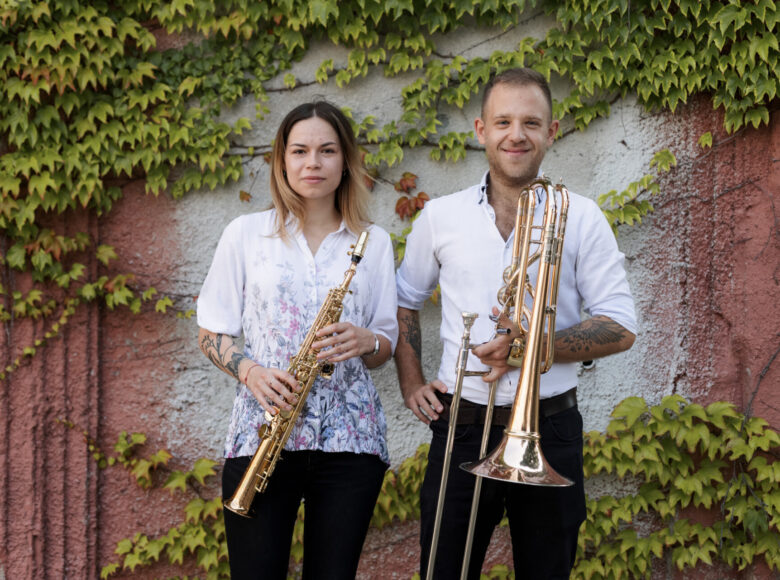 Zdjęcie przedstawia parę młodych muzyków, którzy stoją na tle ściany pokrytej bluszczem. Jest środek dnia. Od lewej strony kobieta obok niej mężczyzna.Mężczyzna jest wyraźnie zadowolony i uśmiechnięty, a kobieta ma odrobinę bardziej poważny wyraz twarzy. Oboje trzymają w rękach instrumenty dęte. Kobieta ma klarnet a mężczyzna puzon.