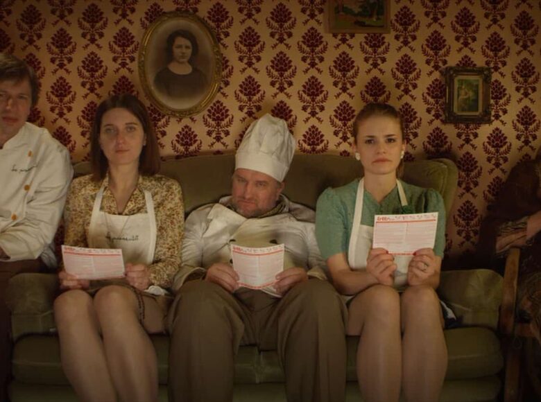 Kadr z filmu. Wnętrze, na kanapie siedzą cztery osoby – dwie kobiety i dwóch mężczyzn, wszyscy są w białych fartuchach, jeden mężczyzna w wysokiej, białej, kucharskiej czapce. Na fotelu obok kanapy siedzi starsza pani, która robi na drutach i się uśmiecha. Osoby z kanapy trzymają w rękach kartki i w skupieniu patrzą na coś przed nimi. W tle ściana z wzorzystą tapetą, na szafce popiersie Matki Boskiej i lampka w abażurze z frędzelkami.