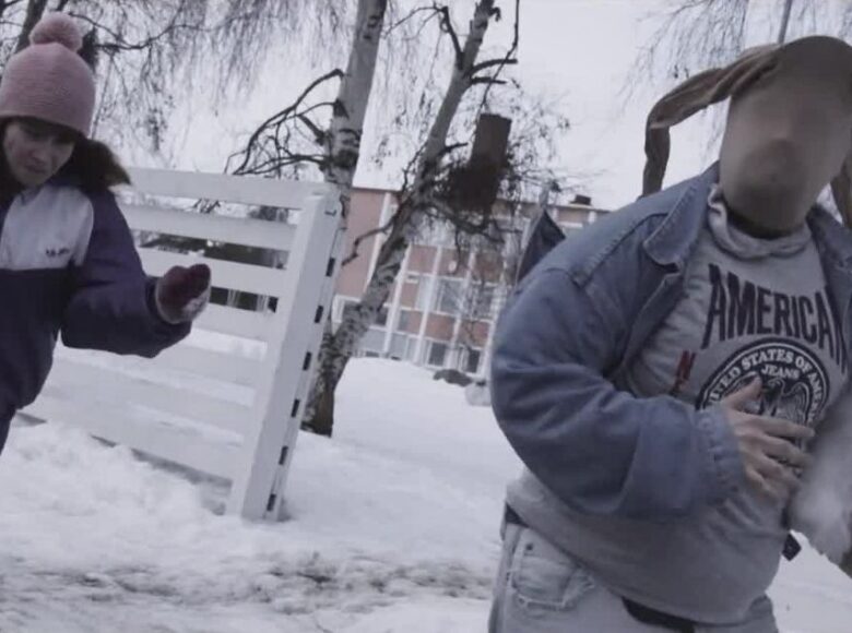 Kadr z filmu. Na zewnątrz, śnieg. Biegną dwie osoby: mężczyzna w dżinsowej kurtce, z rajstopami na głowie i z białym pieskiem pod pachą. Za nim biegnie kobieta w fioletowych dresach i różowej czapce z pomponem.