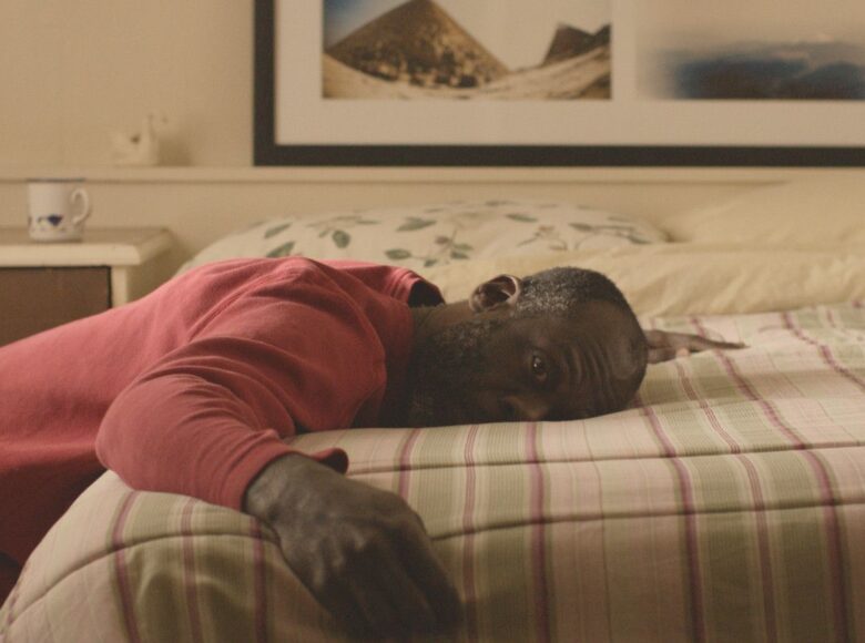 Kadr z filmu. Wnętrze sypialni. Czarnoskóry mężczyzna klęcząc na podłodze głową i tułowiem leży na łóżku. Jest w czerwonej bluzie.