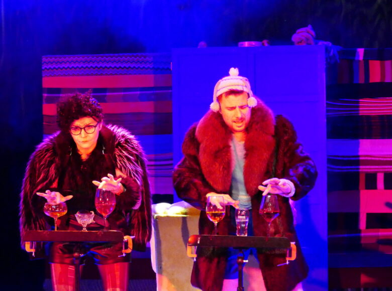 Zdjęcie ze spektaklu "Molowe przypadki". Dwoje aktorów, ubranych w futra stoi przed stolikami, na których znajdują się kieliszki wypełnione płynami. Na scenę pada fioletowe i czerwone światło.