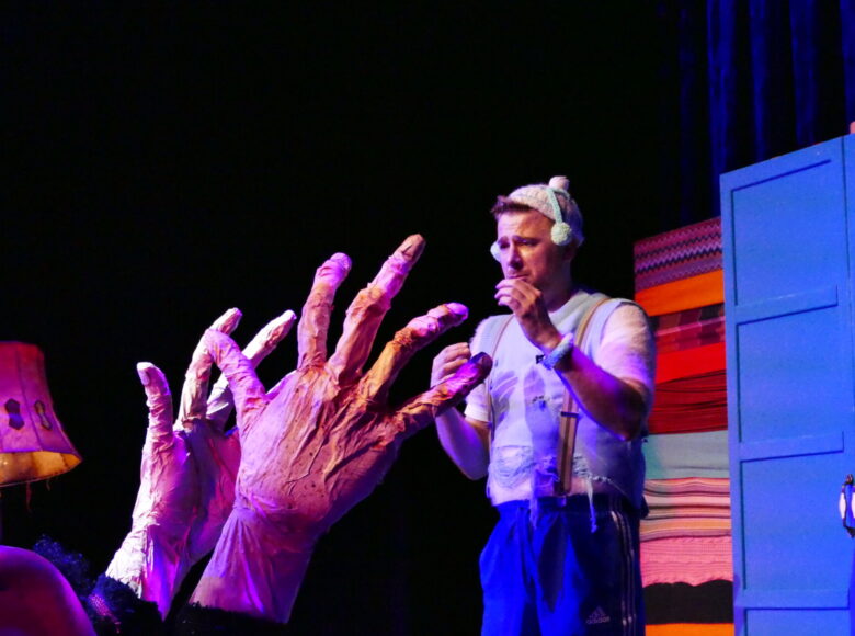 Zdjęcie ze spektaklu "Molowe przypadki". Przedstawia mężczyznę, który stoi na przeciwko wielkich dłoni wystających z czerwonej kanapy. Na scenę pada niebieskie światło.