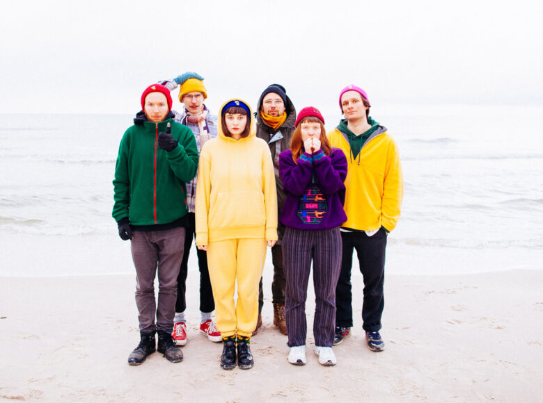 Na zdjeciu grupa 6 osób. Stoją na plaży na brzegu wody. Stoją blisko siebie ubrani w kolorowe ubrania zimowe, kurtki śmieszne czapki, szaliki.
