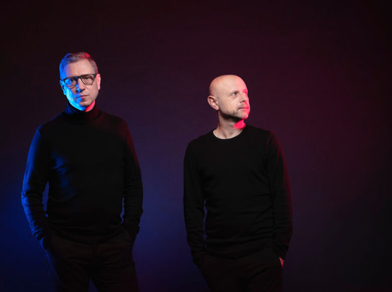 Na fotografi znajduje się dwóch dorosłych panów, którzy stoją w pustej przestrzeni. Obaj są ubrani jednolicie, tak, że zakryte są ich całe ciała oprócz głów. Mają po dwie ręce w kieszeniach. Mężczyzna z lewej strony ma głowę skierowaną w obiektyw. Ma okulary, a jego wyraz twarzy jest poważny. Drugi z panów jest niższy. Jego głowa jest skierowana w lewo. Wygląda na skupionego i zamyślonego.