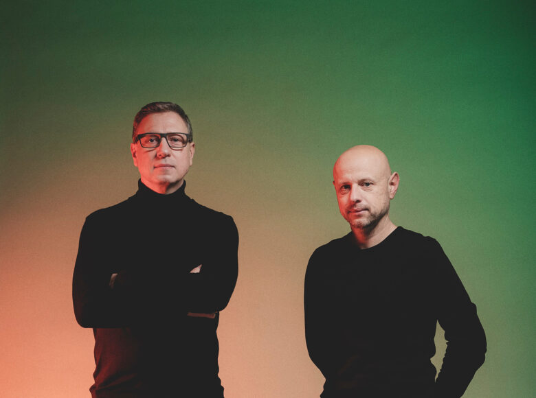 Zdjęcie przedstawia dwóch dorosłych muzyków, którzy występują w pustej przestrzeni. Widać ich tylko do pasa. Mężczyzna po lewej stronie ma okulary a jego ręce są założone i schowane dłonie. Jego postawa jest wyprostowana i z poważną miną. Człowiek stojący obok ma dłonie schowane za plecami. Jest delikatnie pochylony i z lekkim uśmiechem spogląda w obiektyw.