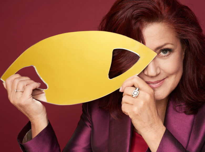 Na zdjęciu znajduje się dojrzała, elegancka kobieta, która trzyma w obu swoich dłoniach lustro o nieregularnym kształcie. Jest to kształt, owalny, lekko spłaszczony z w kierunku końcówek, które się zaostrzają. W środku lustra znajdują się dwa otwory. Przez jeden kobieta przeplata palce prawej dłoni. Drugi otwór przykłada w miejsce prawego oka, którego nie widać, bo przykryte jest włosami. Widocznym okiem kobieta zerka ku górze i delikatnie unosi usta do uśmiechu.