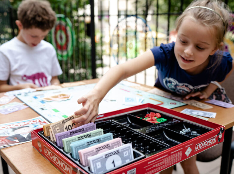 Na zdjęciu dwoje dzieci grających w monopol. Dziewczyna wyciąga rękę i bierze "100zł" z pudełka z grą, chłopiec przegląda karty