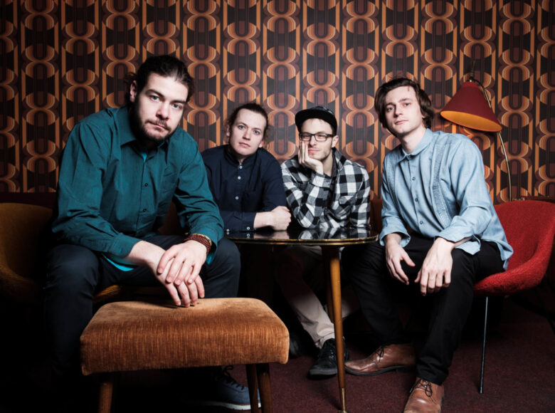 Na pierwszym planie zdjęcia widać dwóch młodych mężczyzn, którzy siedzą na krzesłach. Z lewej strony mężczyzna ma zarost i jest delikatnie zgarbiony. Trzyma łokcie na kolanach, dotykając palcami prawej dłoni o stojący przed nim taboret. Pan po prawej stronie ma delikatnie dłuższe włosy, które zasłaniają mu uszy. Między nimi jest stół, za którym znajduje się druga para młodych muzyków. Mężczyzna po lewej stronie trzyma ręce na stole, natomiast mężczyzna z prawej opiera łokieć na stole i dłonią przytrzymuje brodę. Jako jedyny ma czapkę z daszkiem i okulary. Wszyscy ubrani są w eleganckie koszule.