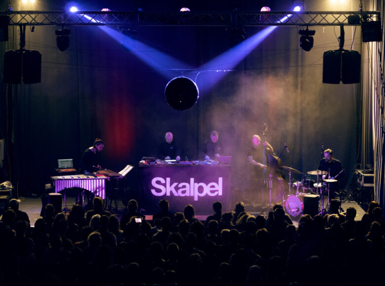 W dolnej części zdjęcia widać liczną grupę ludzi, którzy zwróceni są tyłem do obiektywu. Wszyscy patrzą na scenę, w której centrum stoi stół z napisem „Skalpel”. Za nim jest dwóch mężczyzn grających na instrumentach elektronicznych, a przed nimi są laptopy. Po lewej stronie jest delikatnie zgarbiony mężczyzna, który trzyma w rękach po dwie pałki i gra na dużych cymbałach. Z prawej strony jest jeszcze dwóch mężczyzn. Pierwszy gra na kontrabasie a drugi na perkusji.