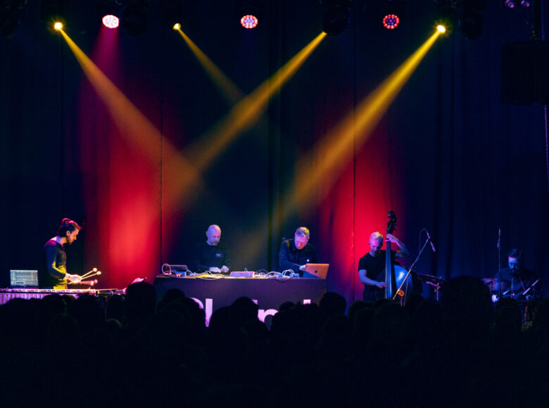 Zdjęcie przedstawia występujących na scenie artystów. Po lewej stronie jest delikatnie zgarbiony mężczyzna, który trzyma w rękach po dwie pałki i gra na dużych cymbałach. W środkowej części sceny znajduje się dwóch panów, którzy stoją za konsolą i używając między innymi laptopów, tworzą muzykę elektroniczną. Po prawej stronie od konsoli stoi kontrabasista a obok niego siedzi perkusista. Każdy z muzyków jest skupiony i zaangażowany w koncert.