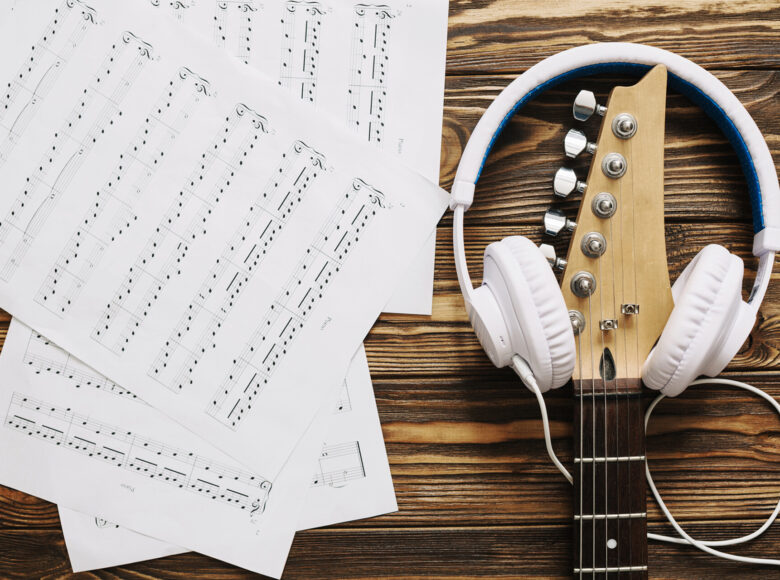 na drewnianym stole po lewej stronie leżą rozrzucone kartki z nutami, po prawej leży gryf od stary i założone na niego duże białe słuchawki.