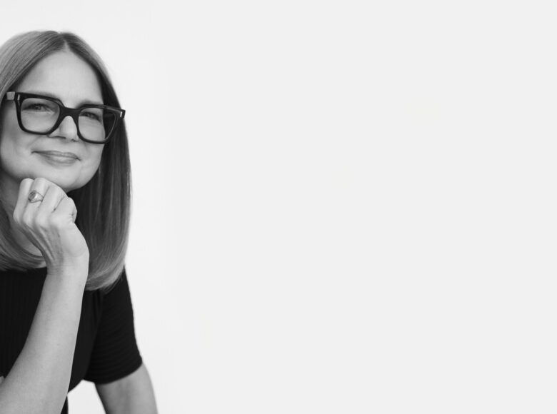 Fotografia czarno biała. Na białym tle zbliżenie na twarz i ramiona kobiety w okularach i w czarnej bluzce.